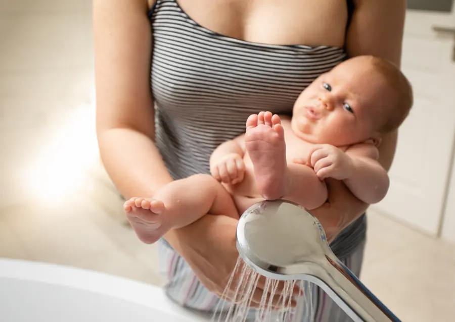 Mutter hält ihr nacktes Baby im Badezimmer sanft unter den warmen Wasserstrahl