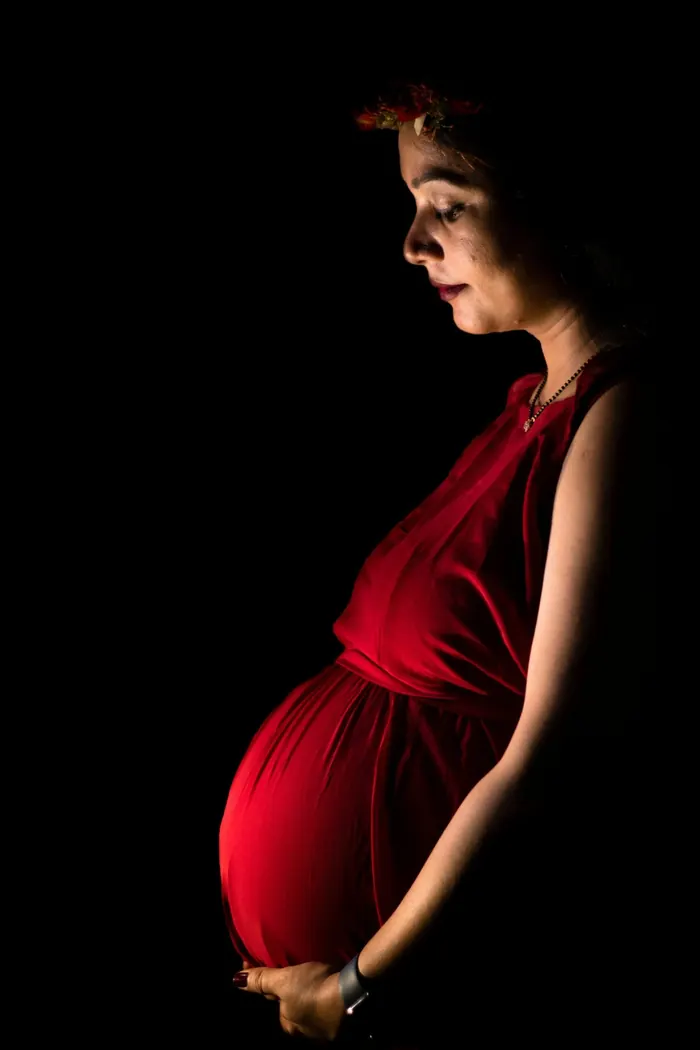Schwangere im roten Kleid steht im warmen Licht vor dunklem Hintergrund und hält den Bauch